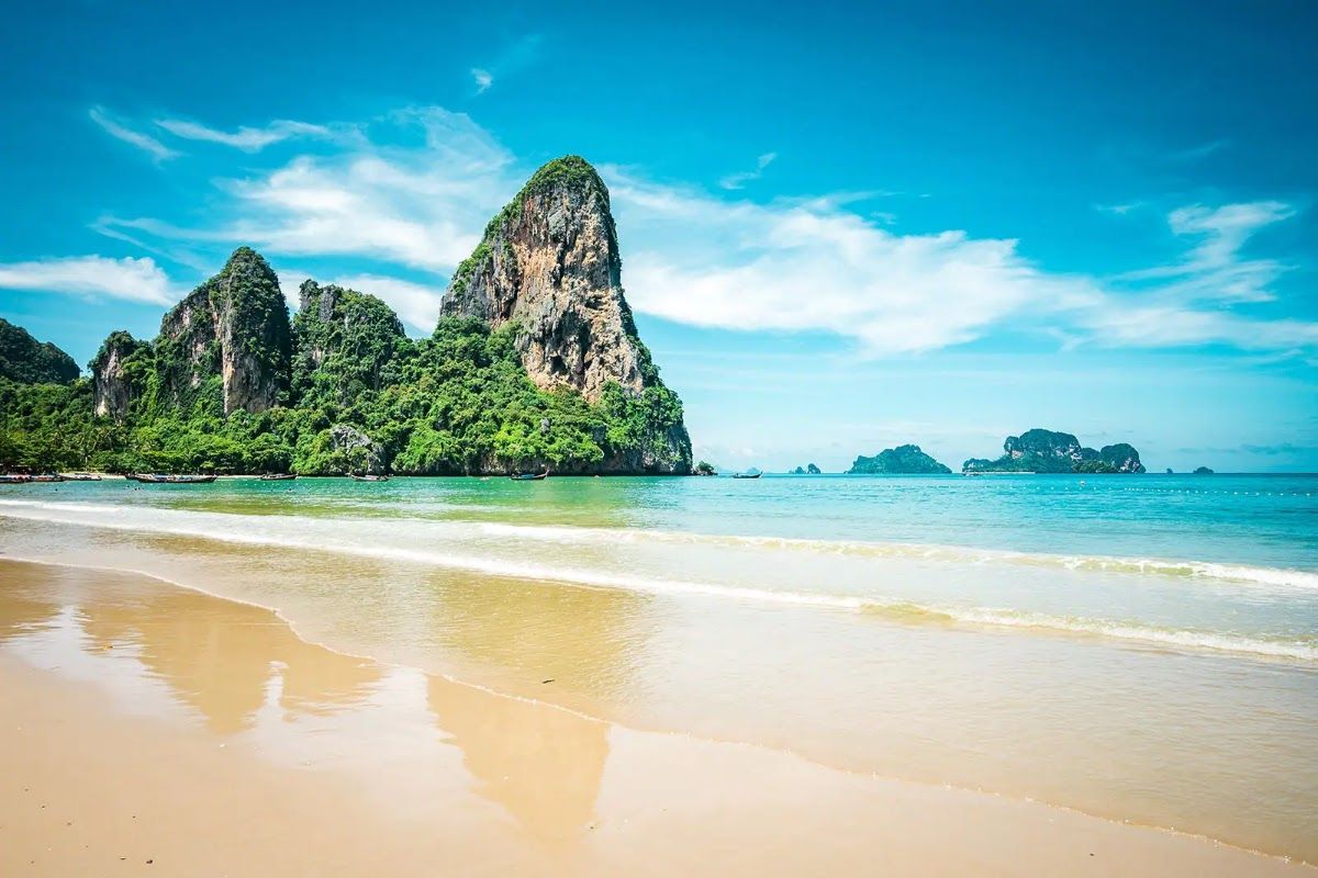 Railay Beach in Krabi with a tall limestone karst cliff rising behind pale sand and turquoise water